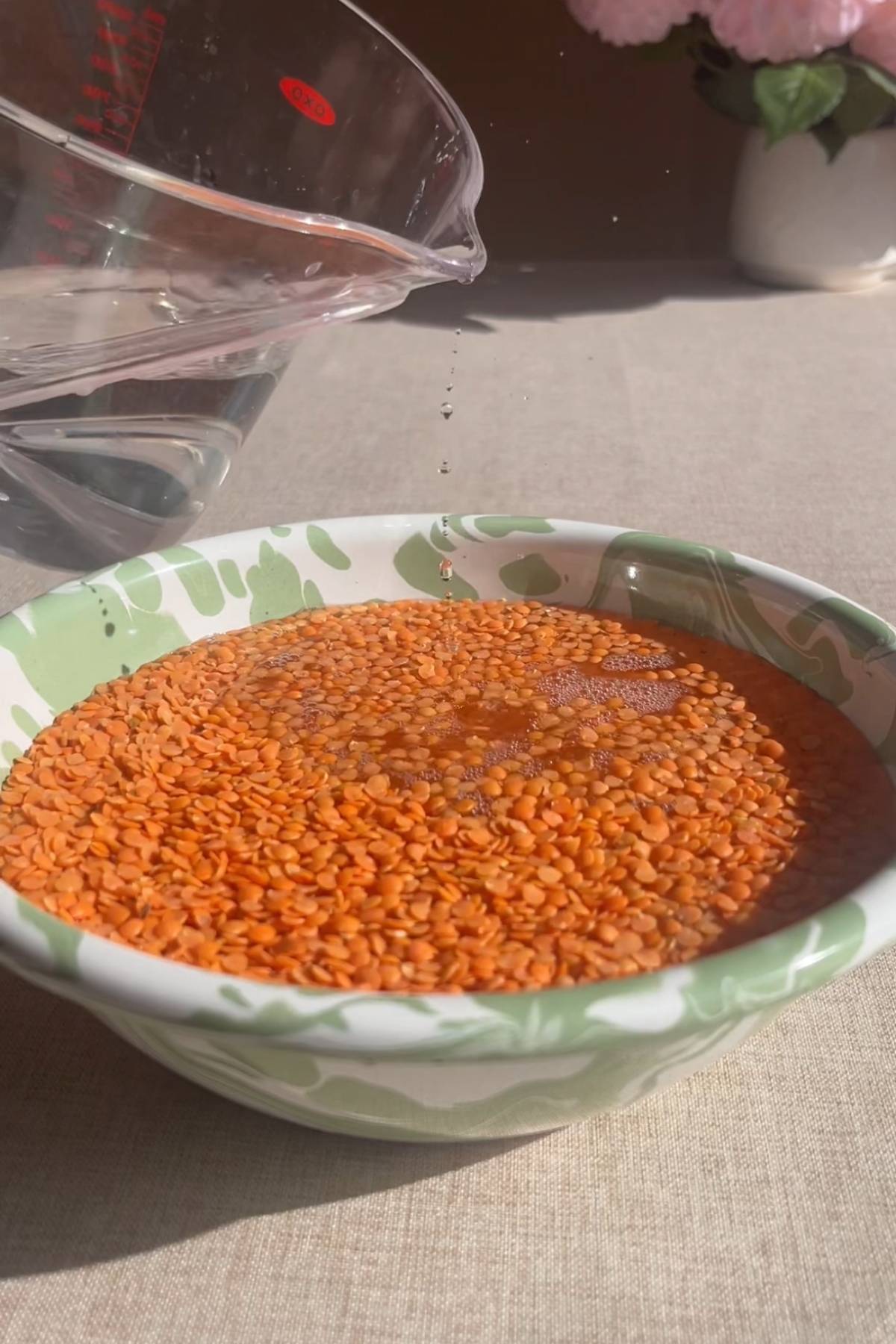 Water being poured from a measuring jug over red lentils in a patterned bowl.