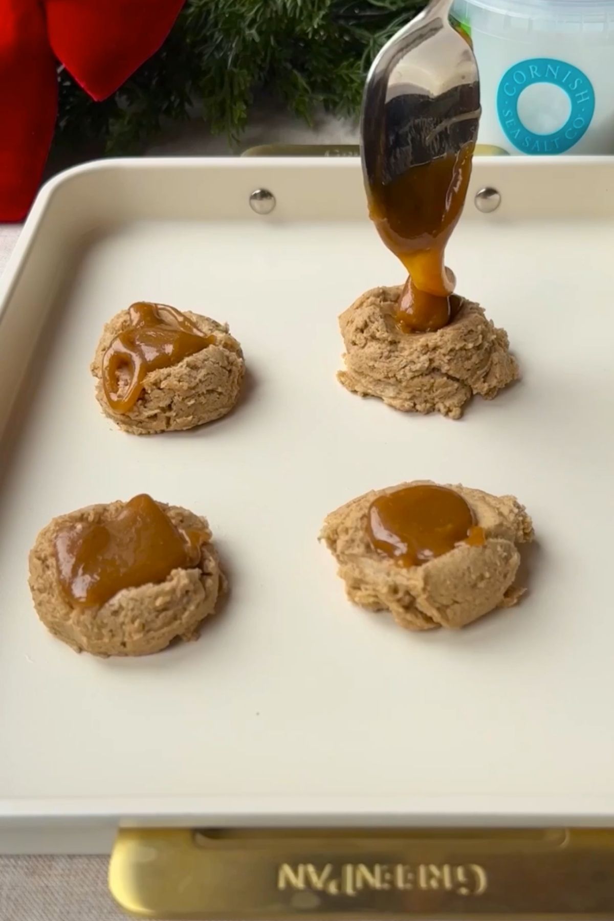 A spoon dropping warm caramel into indents on unbaked cookie dough balls on a tray.
