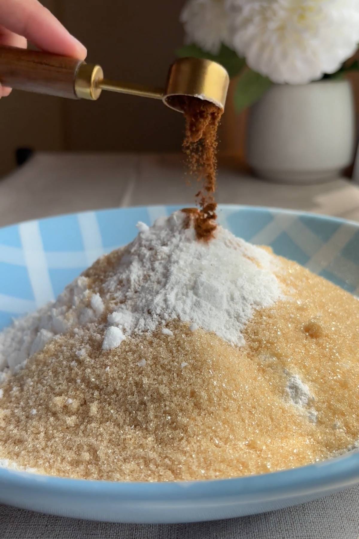 Cinnamon being sprinkled from a gold measuring spoon over sugar and dry ingredients in a bowl.