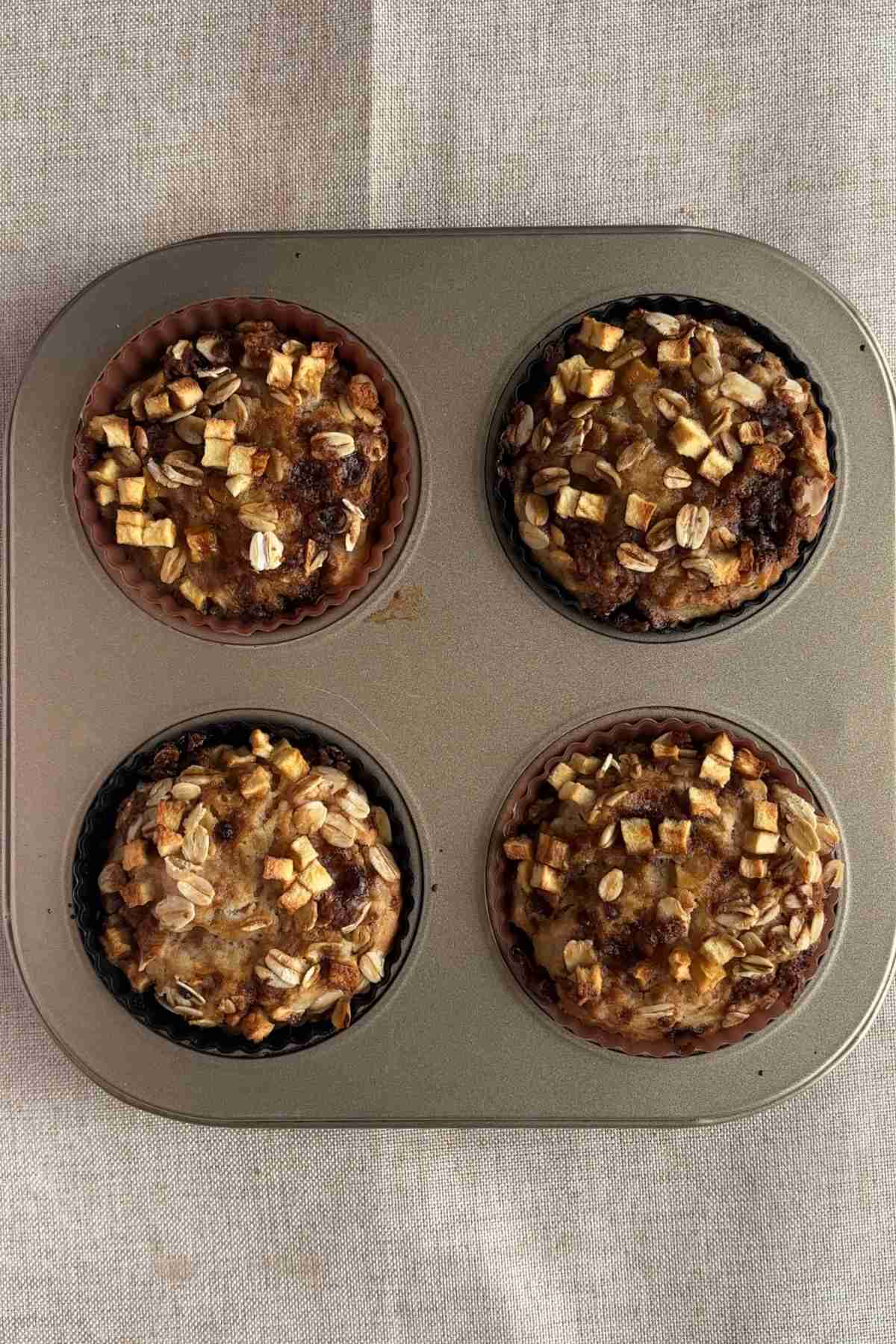 Four baked muffins with oats and small apple pieces on top in a muffin tray.