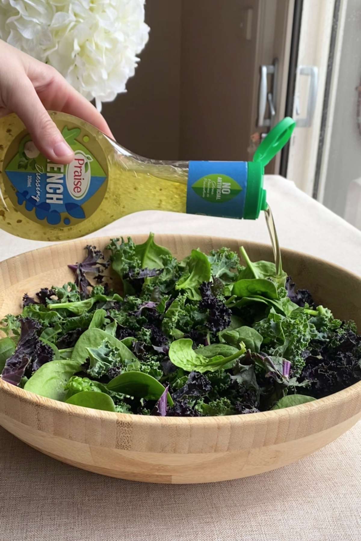 Hand pouring dressing over fresh kale and spinach in a wooden bowl.