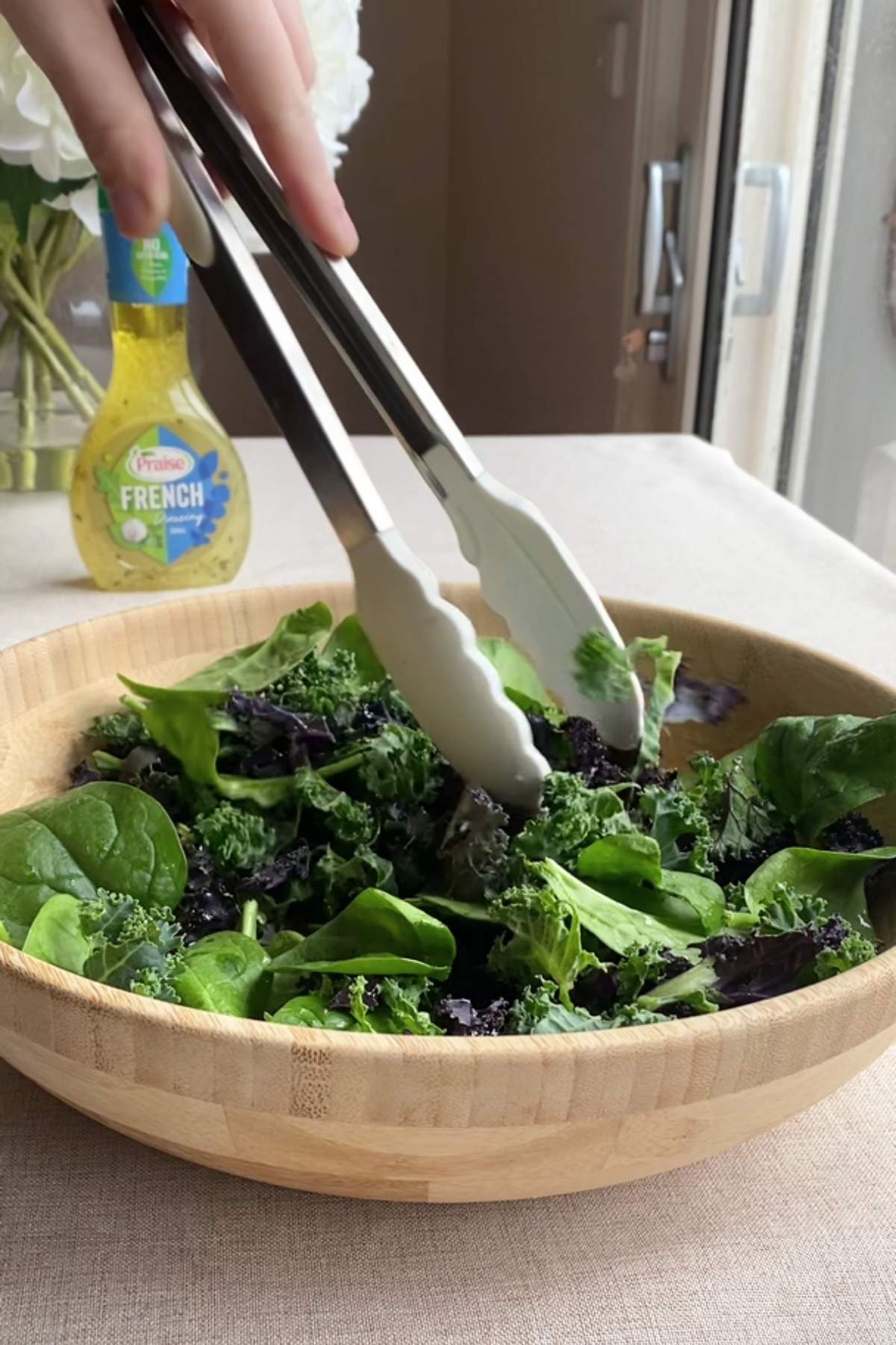 Tongs tossing fresh kale and spinach leaves in a wooden bowl.