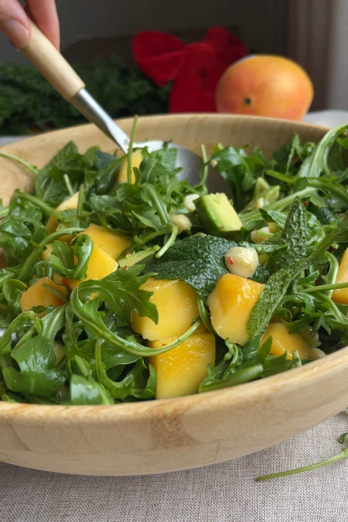 Hand stirring arugula salad with mango and avocado in a wooden bowl.