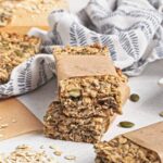 Stacked oat and seed granola bars wrapped in parchment, surrounded by loose oats and pumpkin seeds.