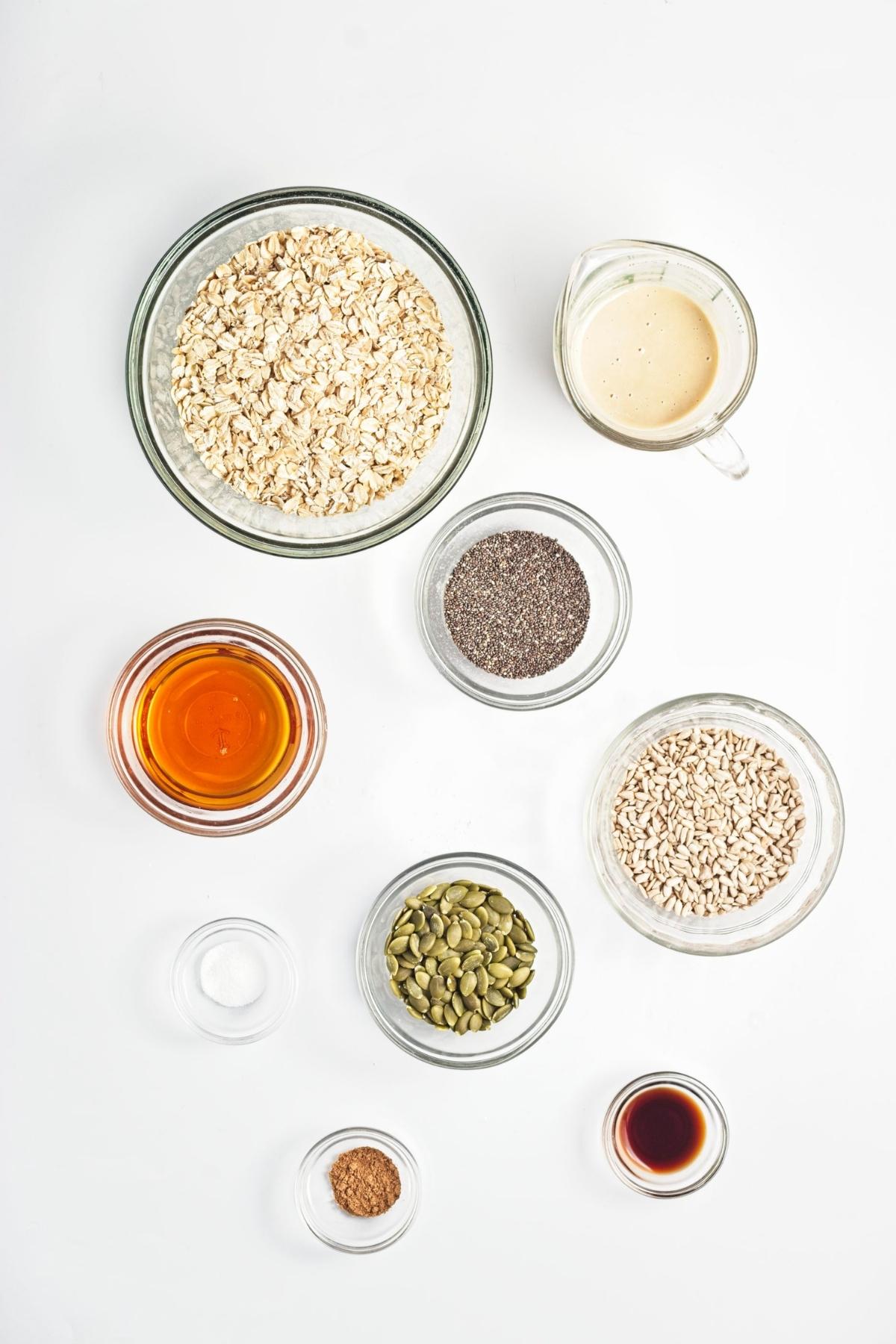 Overhead of oats, seeds, tahini, maple syrup, and spices arranged in small glass bowls.