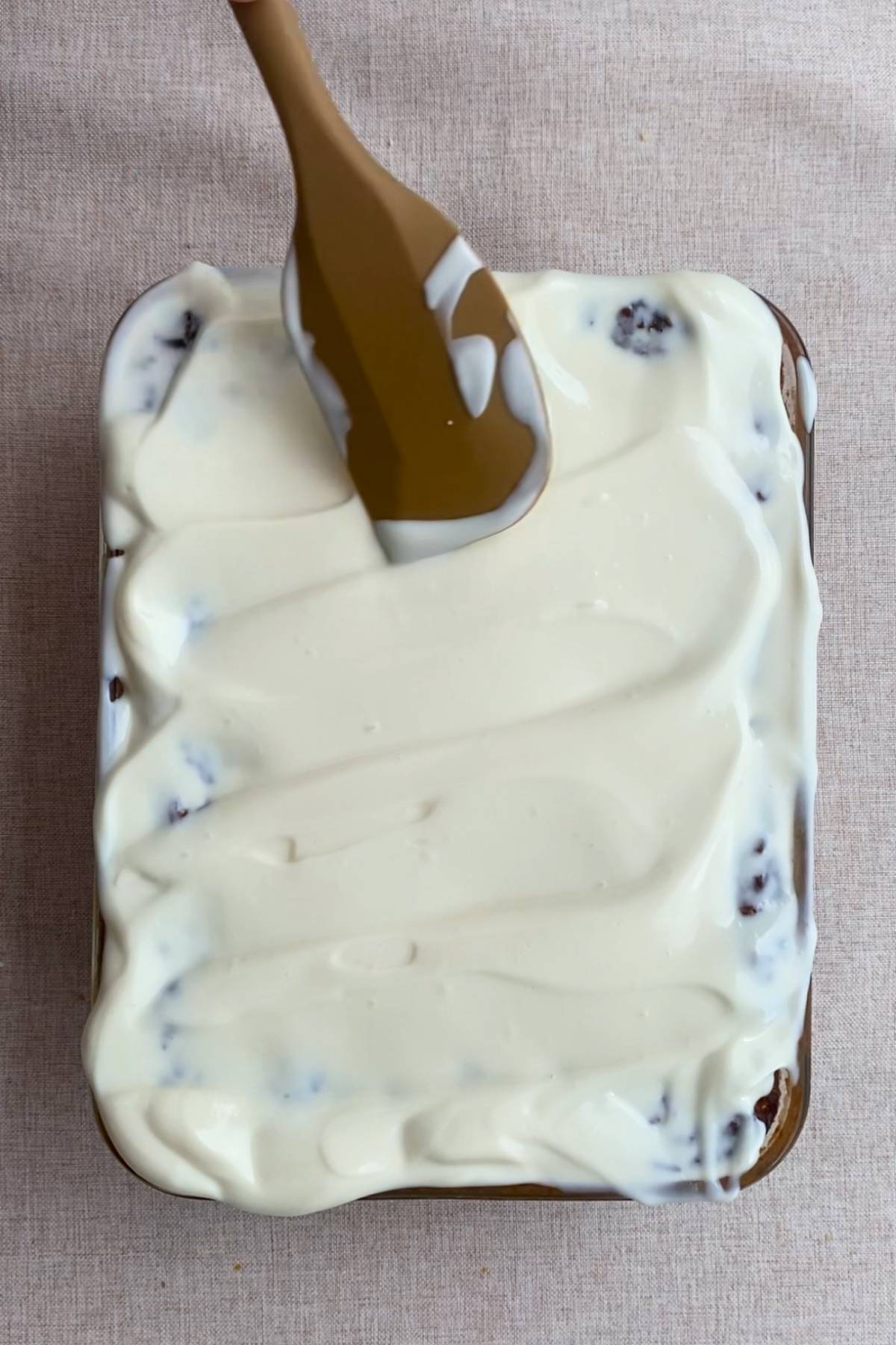 Spreading creamy white frosting evenly over a baked oat mixture with a brown spatula.