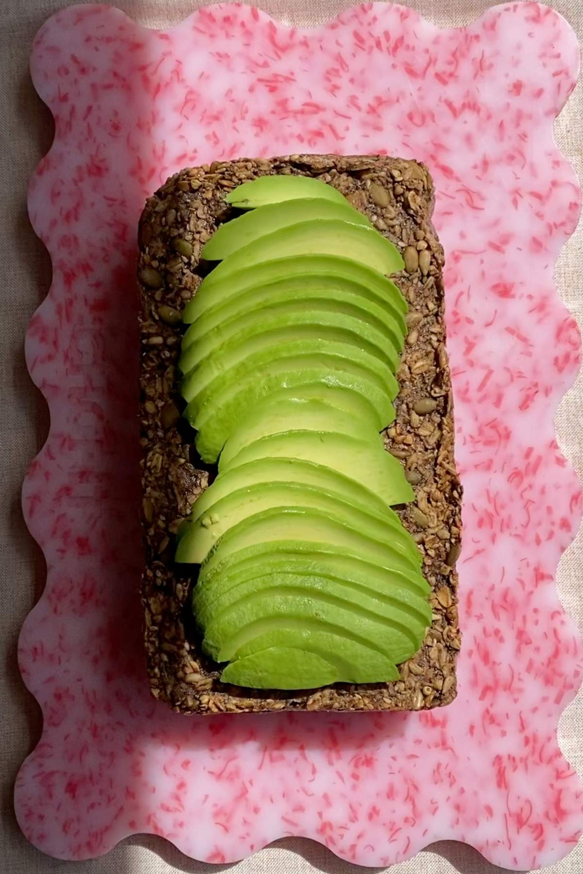 Whole seeded loaf topped with sliced avocado on a pink serving board.