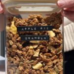Clear container filled with chunky apple pie granola, showing oats, pepitas, pecans, and dried apple pieces.
