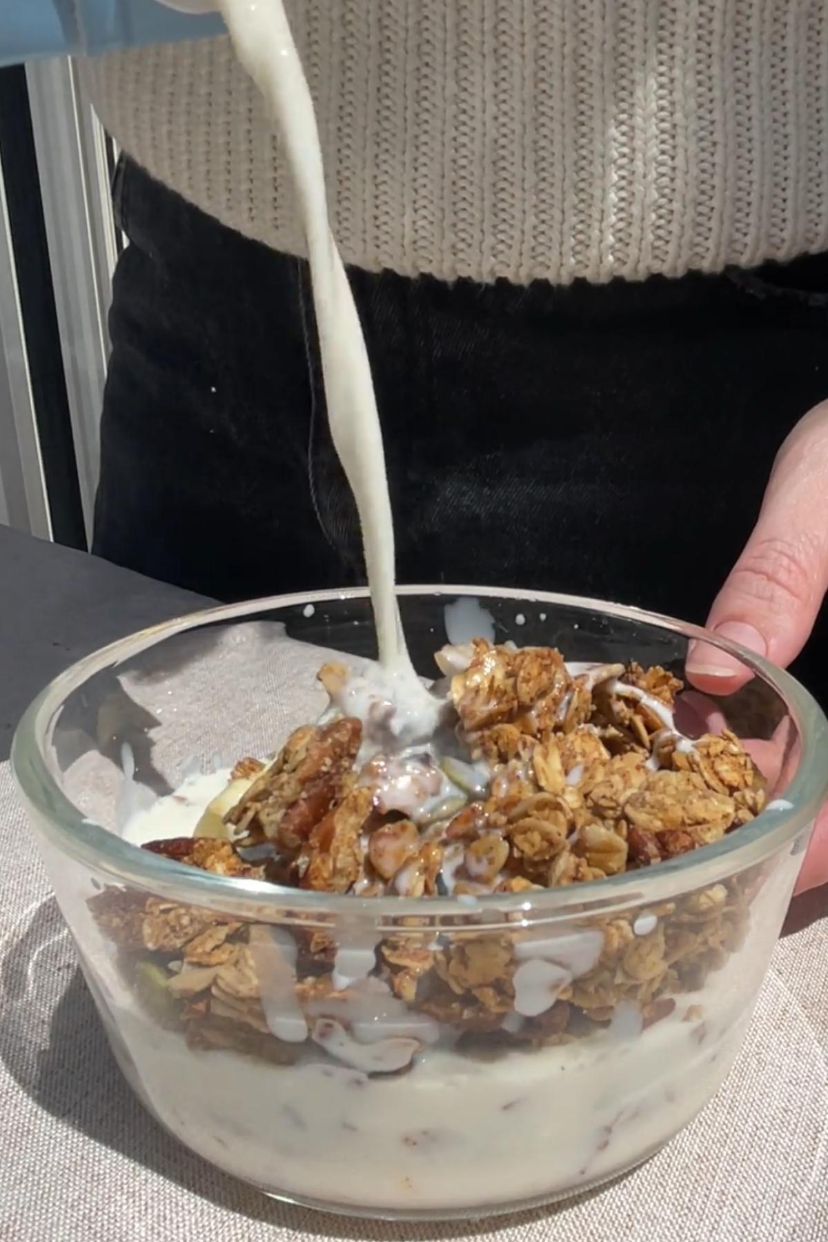 Milk being poured over apple pie granola in a glass bowl, soaking crunchy oat and nut clusters.