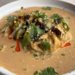 Close-up of baked dumplings resting in thick peanut coconut curry sauce, garnished with fresh coriander, sesame seeds, and chilli oil.
