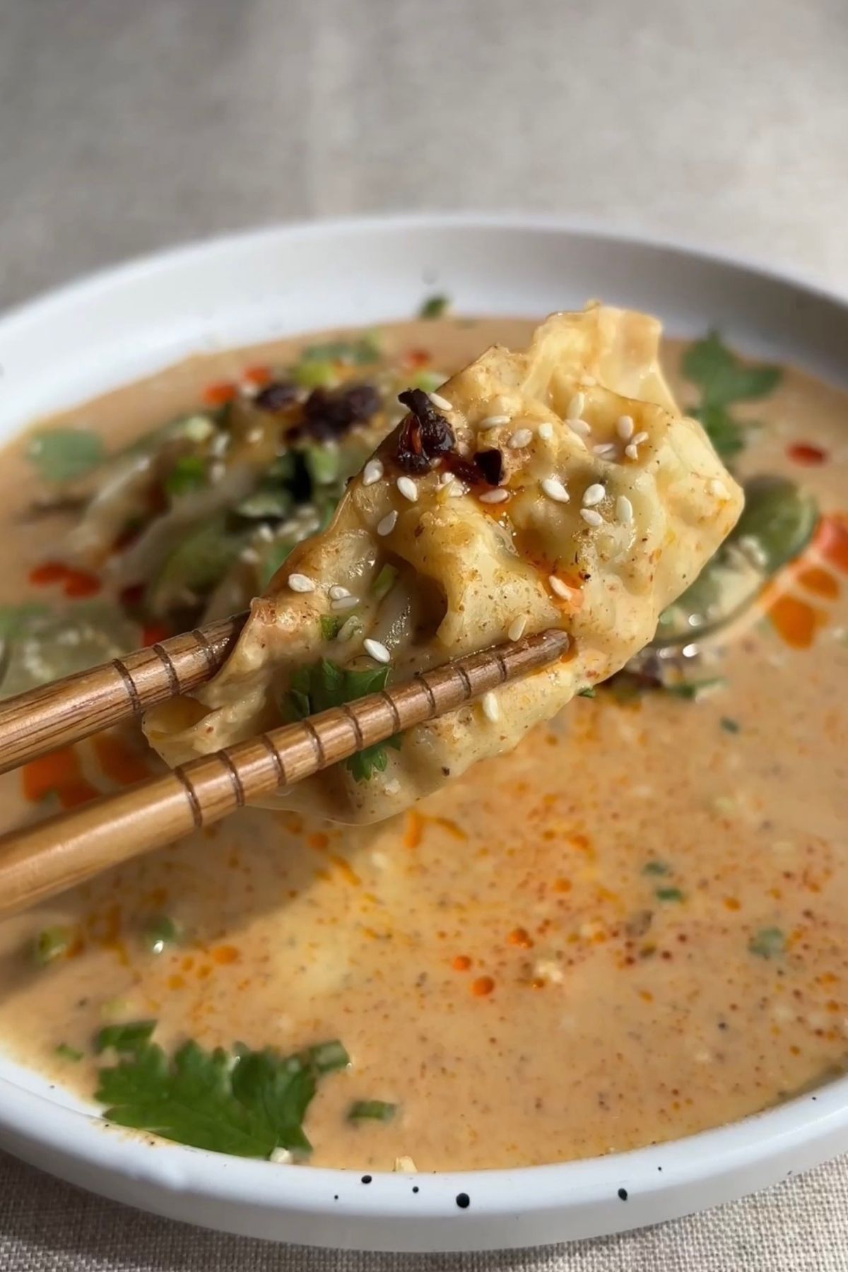 Chopsticks lifting a baked dumpling coated in creamy curry sauce with sesame seeds and chilli oil, showing the soft wrapper and filling.