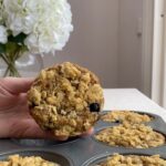 Hand holding a blueberry breakfast muffin with oat streusel topping above a muffin tin.