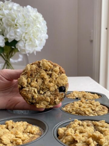 Hand holding a blueberry breakfast muffin with oat streusel topping above a muffin tin.