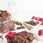 Chocolate granola clusters served in milk with sliced strawberries and a spoon on a white plate.