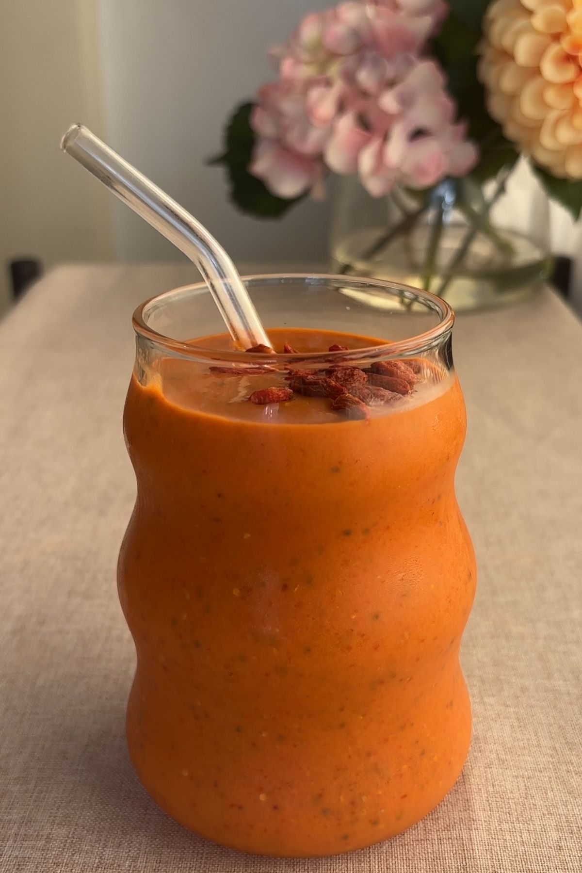 Glass of thick orange smoothie with soaked goji berries and a clear glass straw.