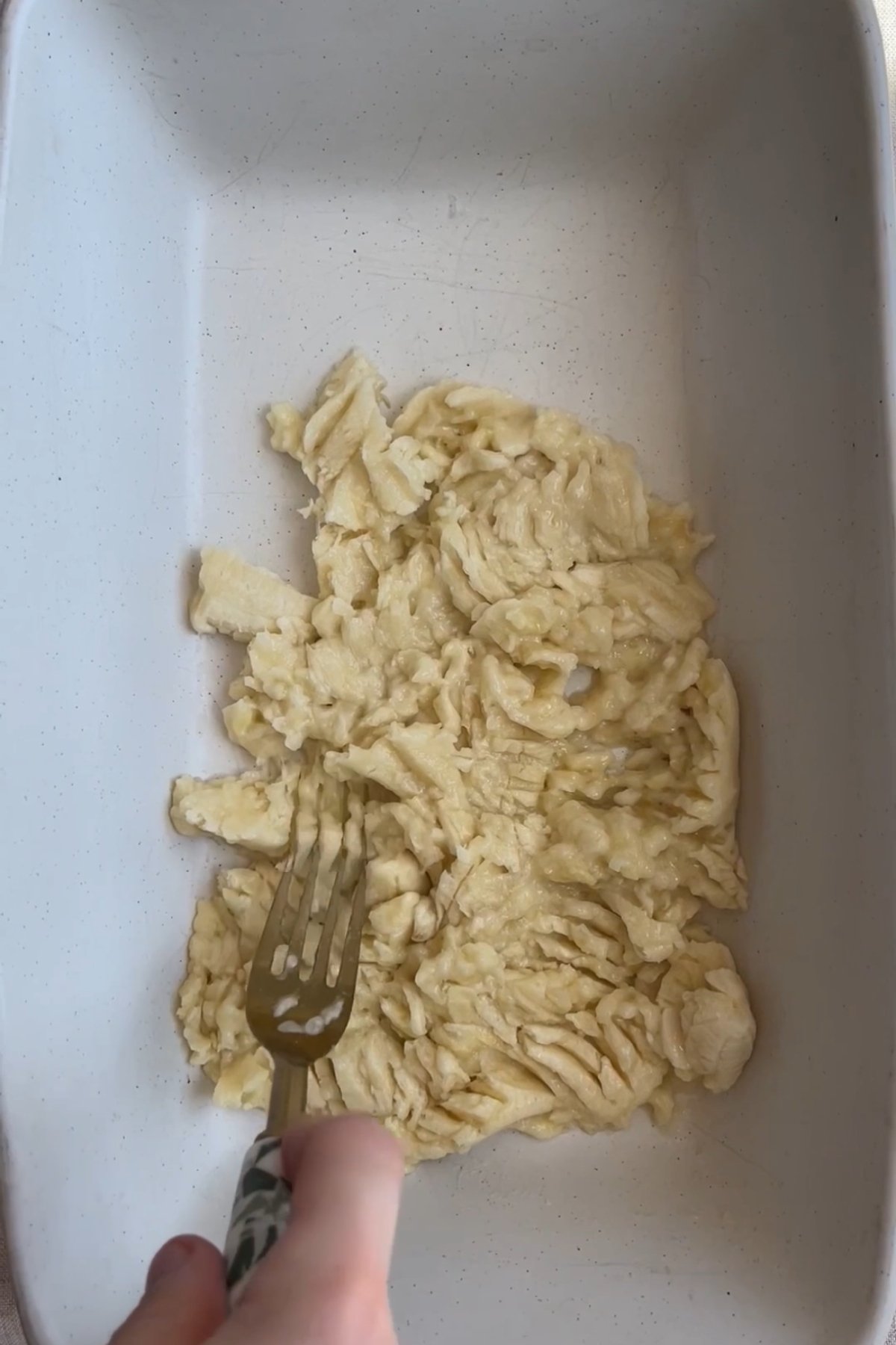 Mashed ripe banana in a baking dish, being pressed with a fork until smooth.