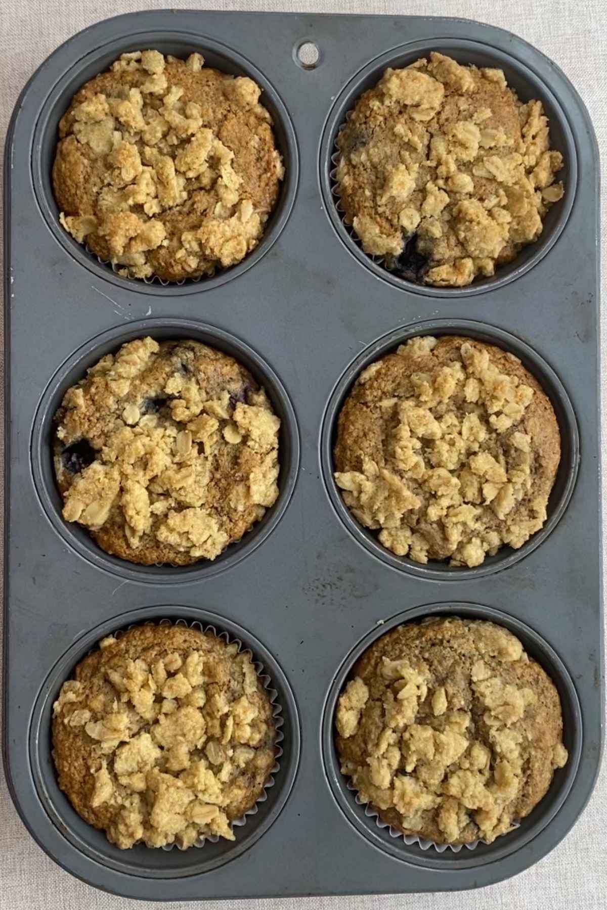 Fully baked blueberry breakfast muffins with golden oat streusel topping in a lined muffin tin, fresh from the oven.