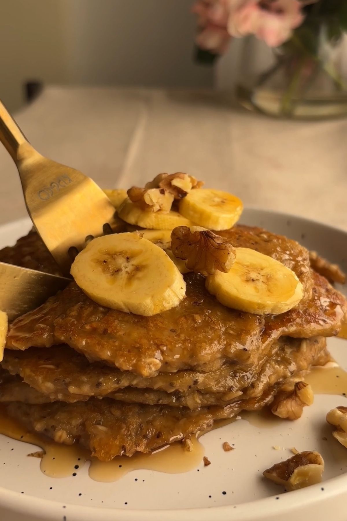 Pancakes topped with banana slices and walnuts being cut with a fork, drizzled with syrup.