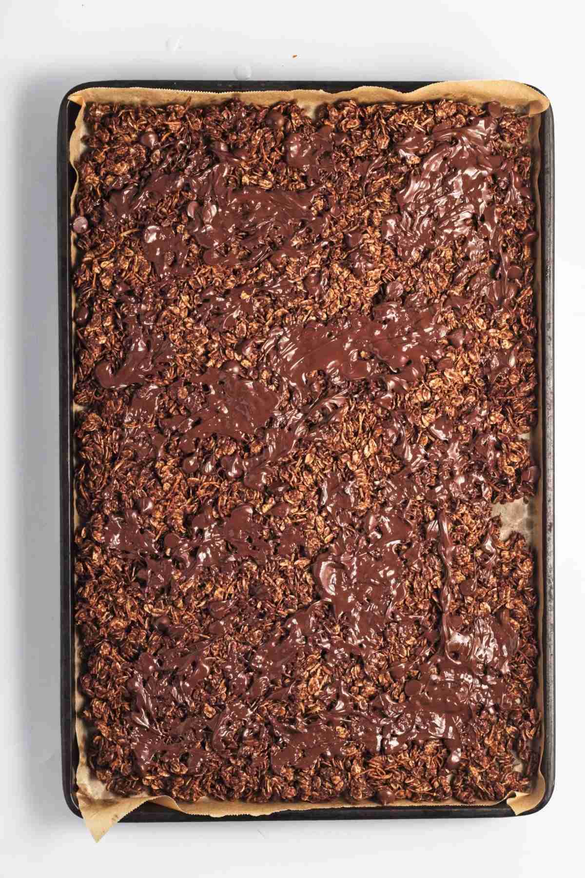 Melted chocolate swirled over baked chocolate granola on a lined baking tray.