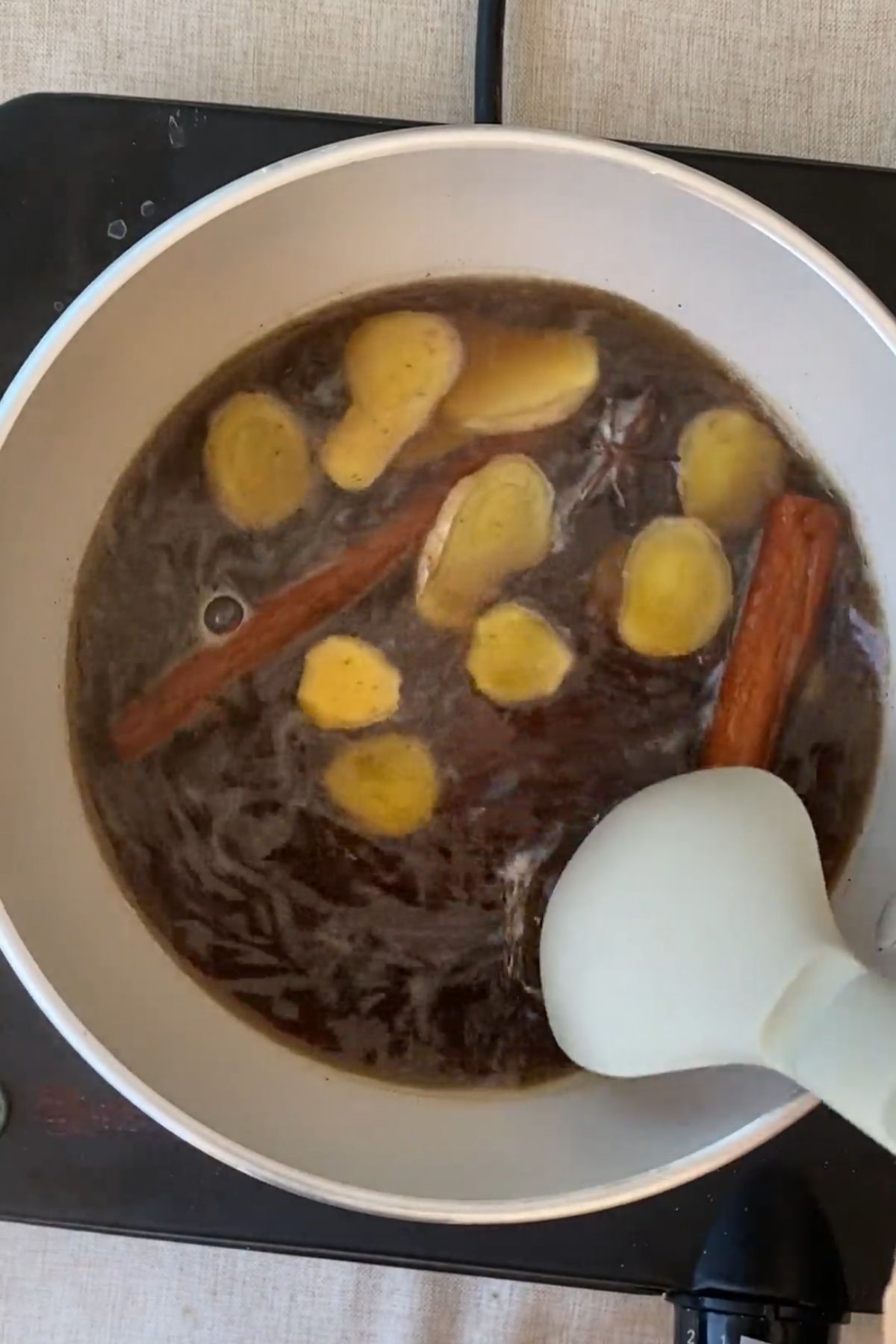 Stirring ginger slices and cinnamon sticks simmering in dark syrup in a saucepan.