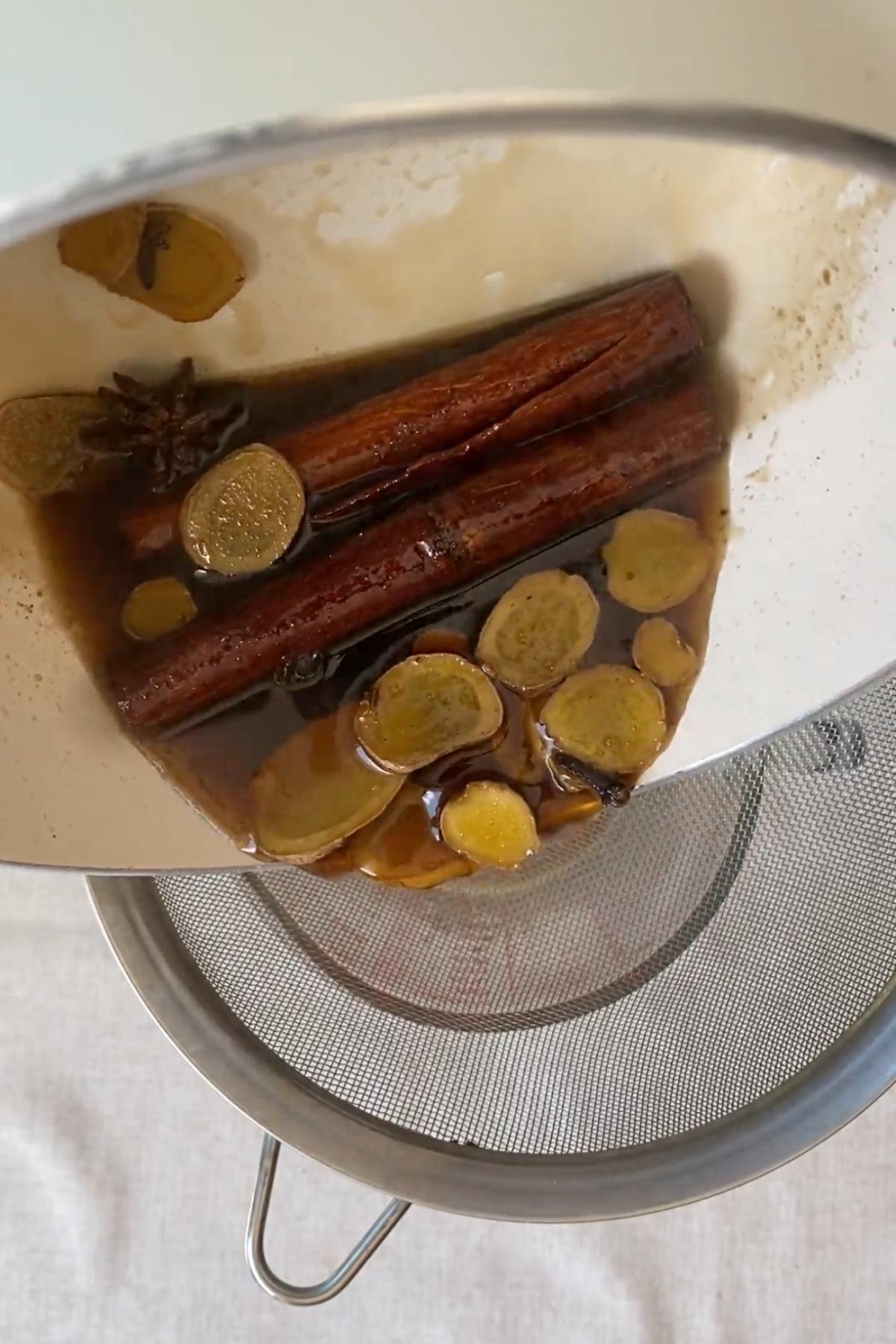 Spiced syrup poured through a fine mesh sieve with ginger and cinnamon visible.