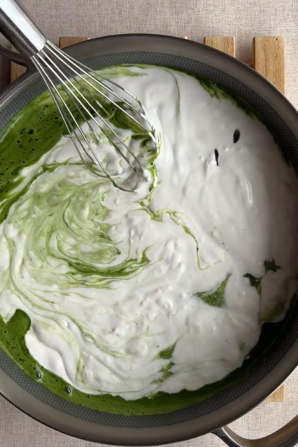 White cream being whisked into a green matcha mixture in a pan.
