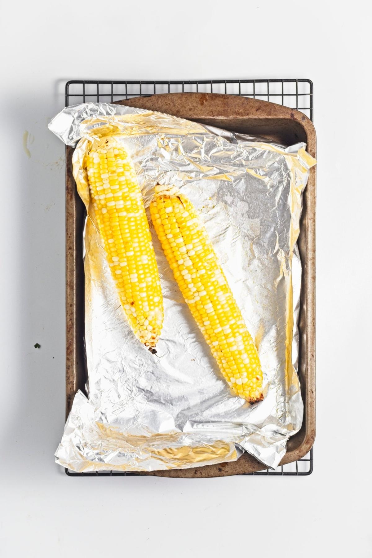 Two whole corn cobs roasting on a foil-lined baking tray.