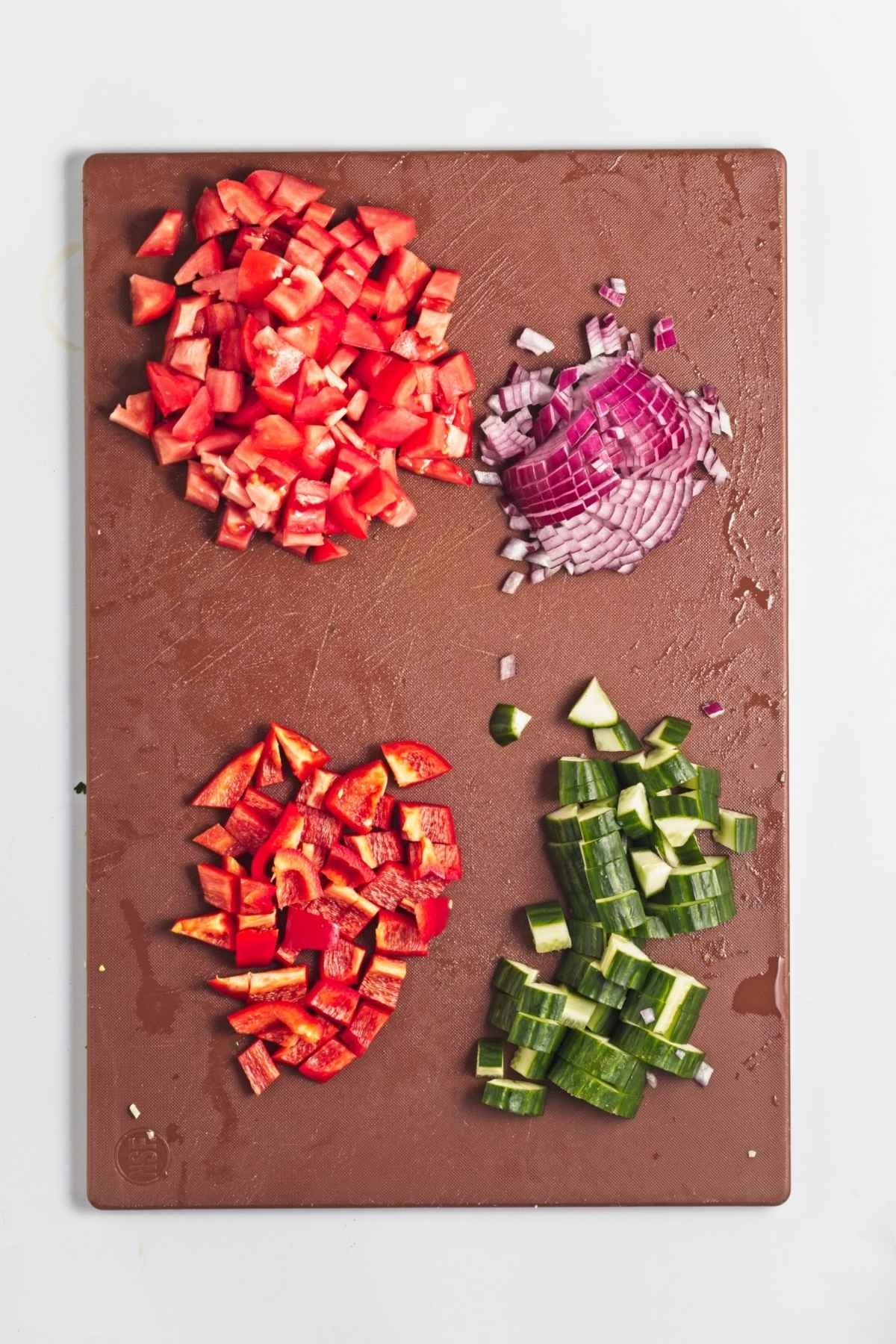 Chopped tomatoes, cucumber, red onion, and red bell pepper arranged on a cutting board.
