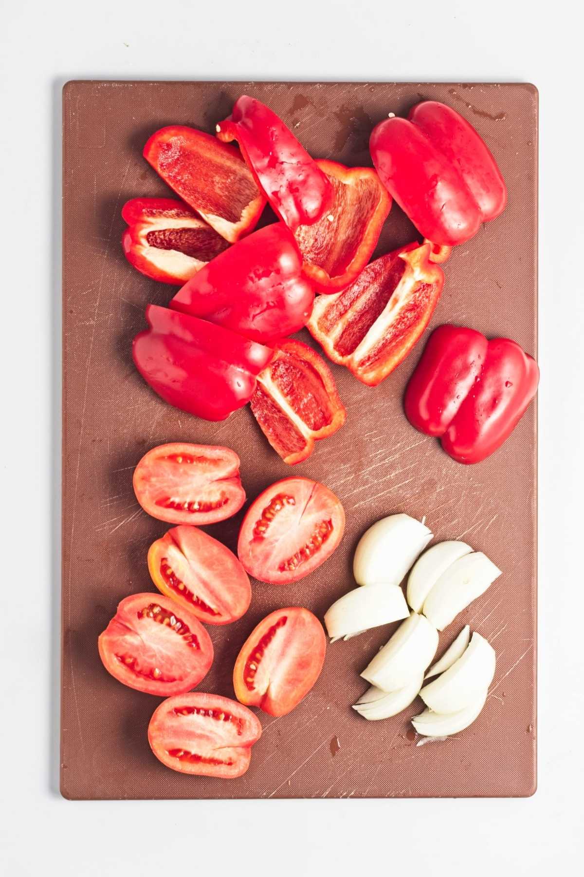 Red bell peppers, tomatoes, onion, garlic, and celeriac chopped on a cutting board.