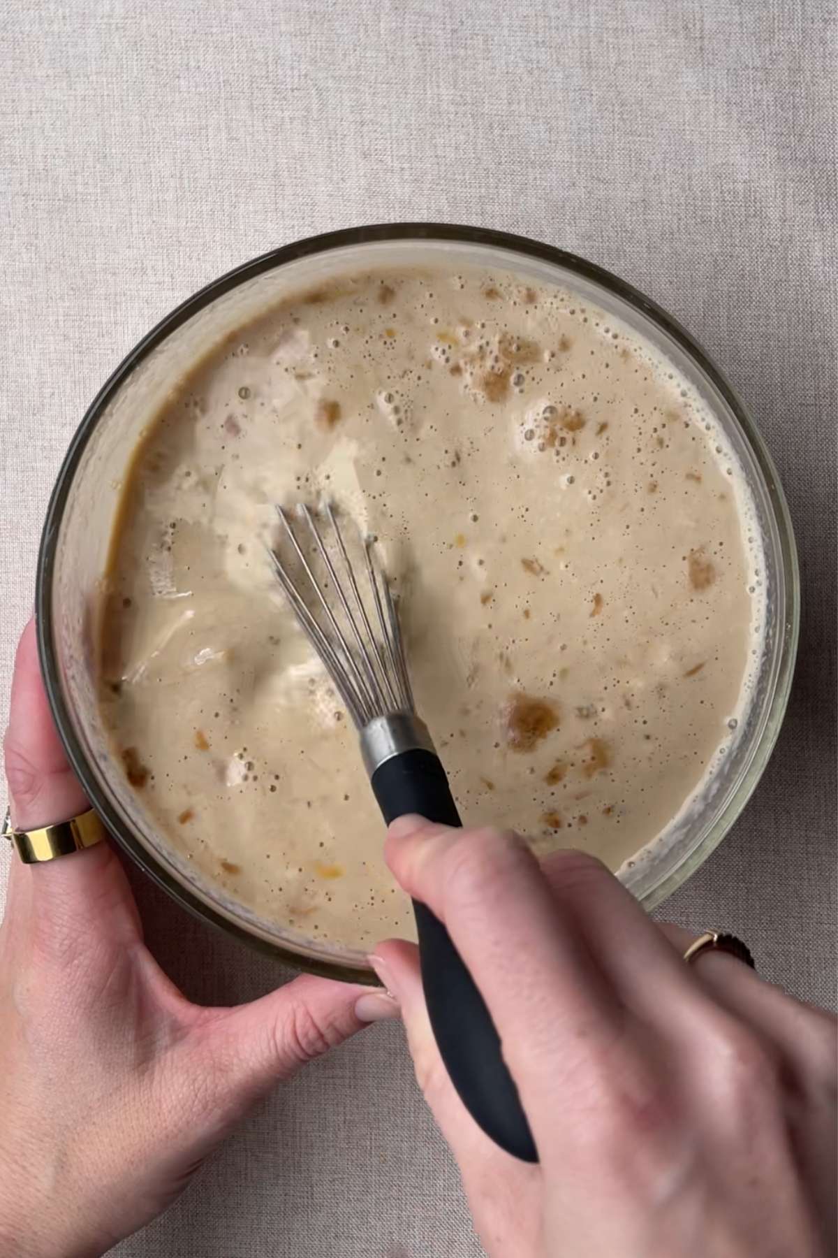 Hands whisking a creamy liquid mixture in a glass bowl.