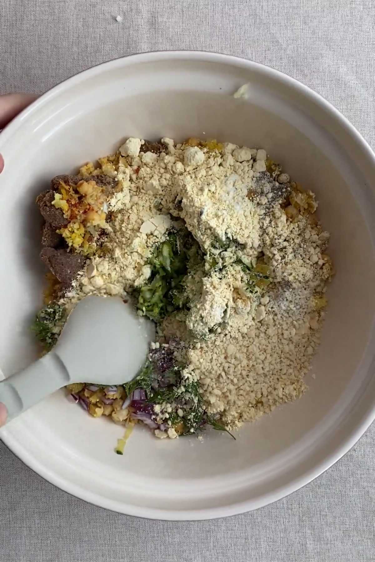 Bowl of fritter mixture with chickpea flour, herbs, and zucchini being mixed.