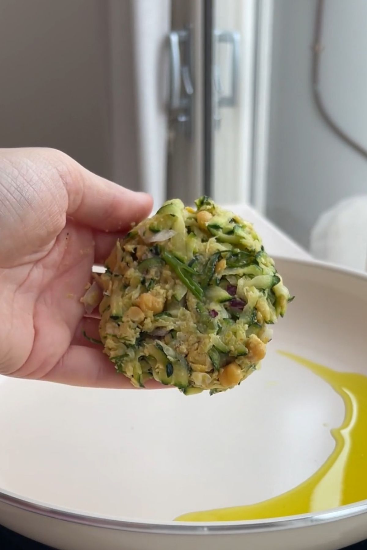 Shaped zucchini chickpea fritter mixture held in a hand above a pan.