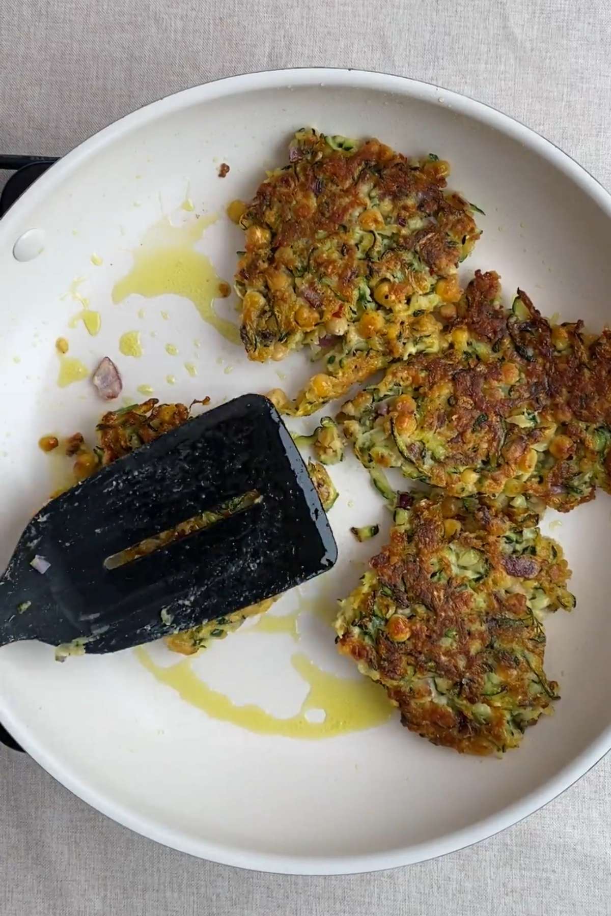 Zucchini chickpea fritters cooking in a skillet with oil and a spatula.