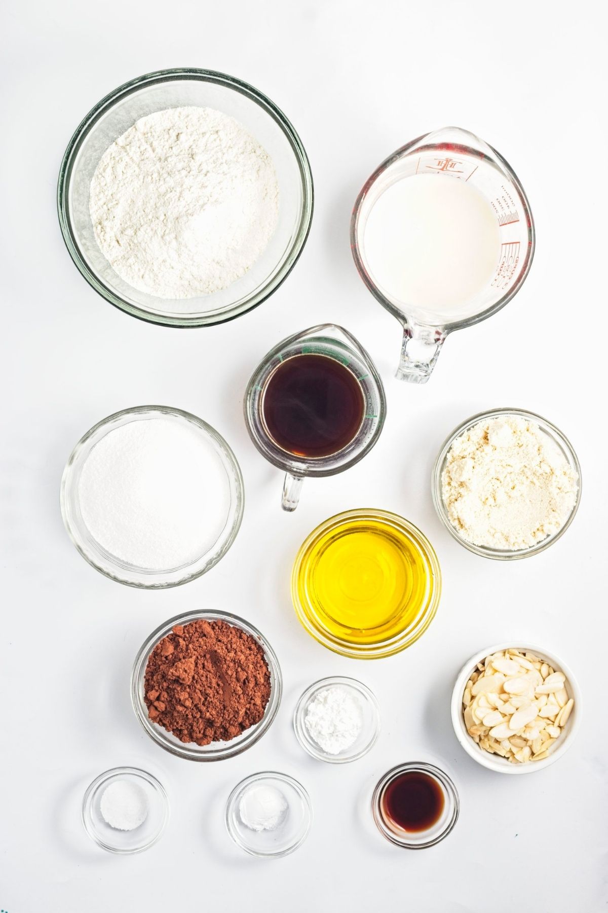 Overhead view of baking ingredients including flour, cocoa powder, sugar, olive oil, coffee, milk, and sliced almonds.