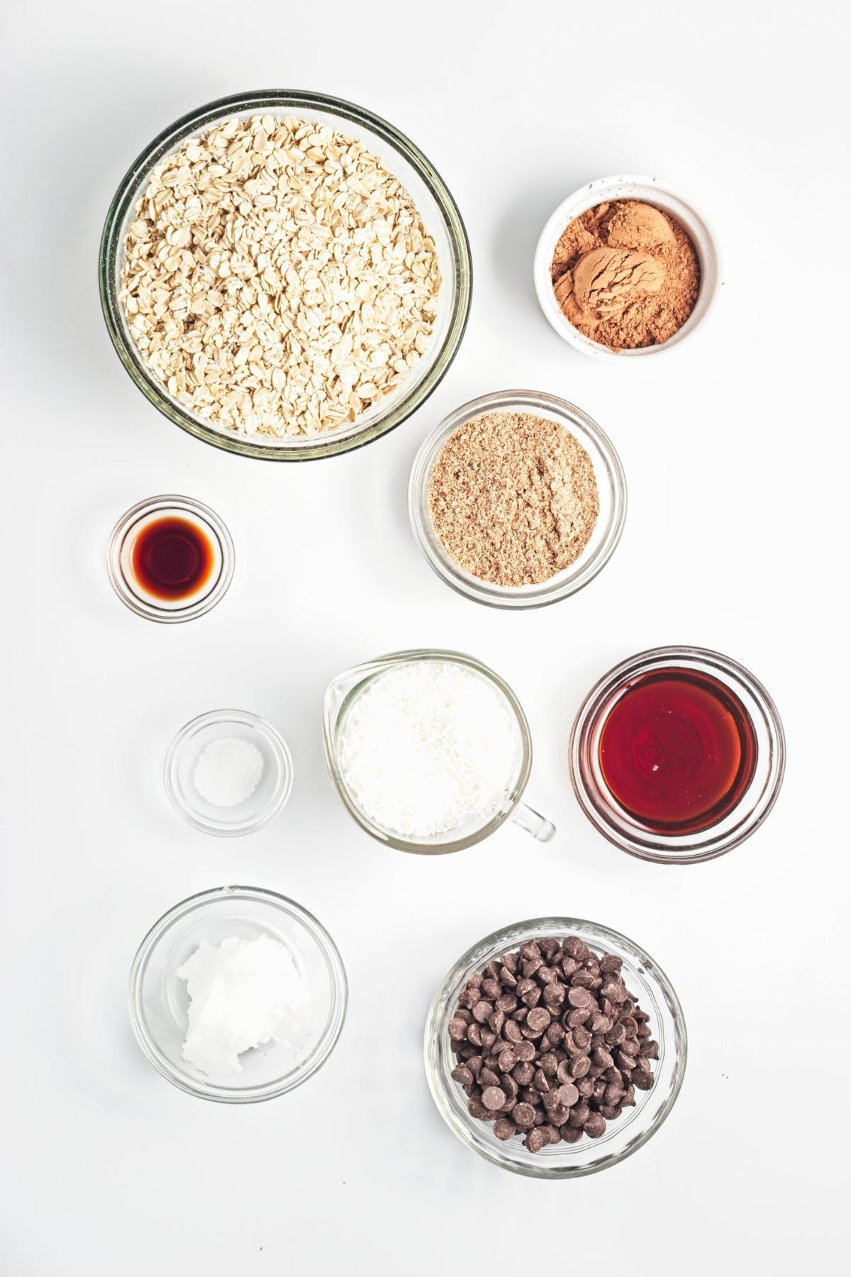 Bowls of rolled oats, cocoa powder, ground flaxseed, shredded coconut, maple syrup, coconut oil, vanilla, salt, and chocolate chips.