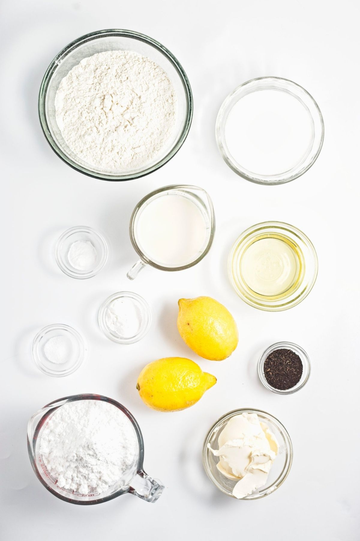 Measured baking ingredients arranged on a white surface, including flour, oat milk, oil, lemons, tea leaves, and vegan butter.