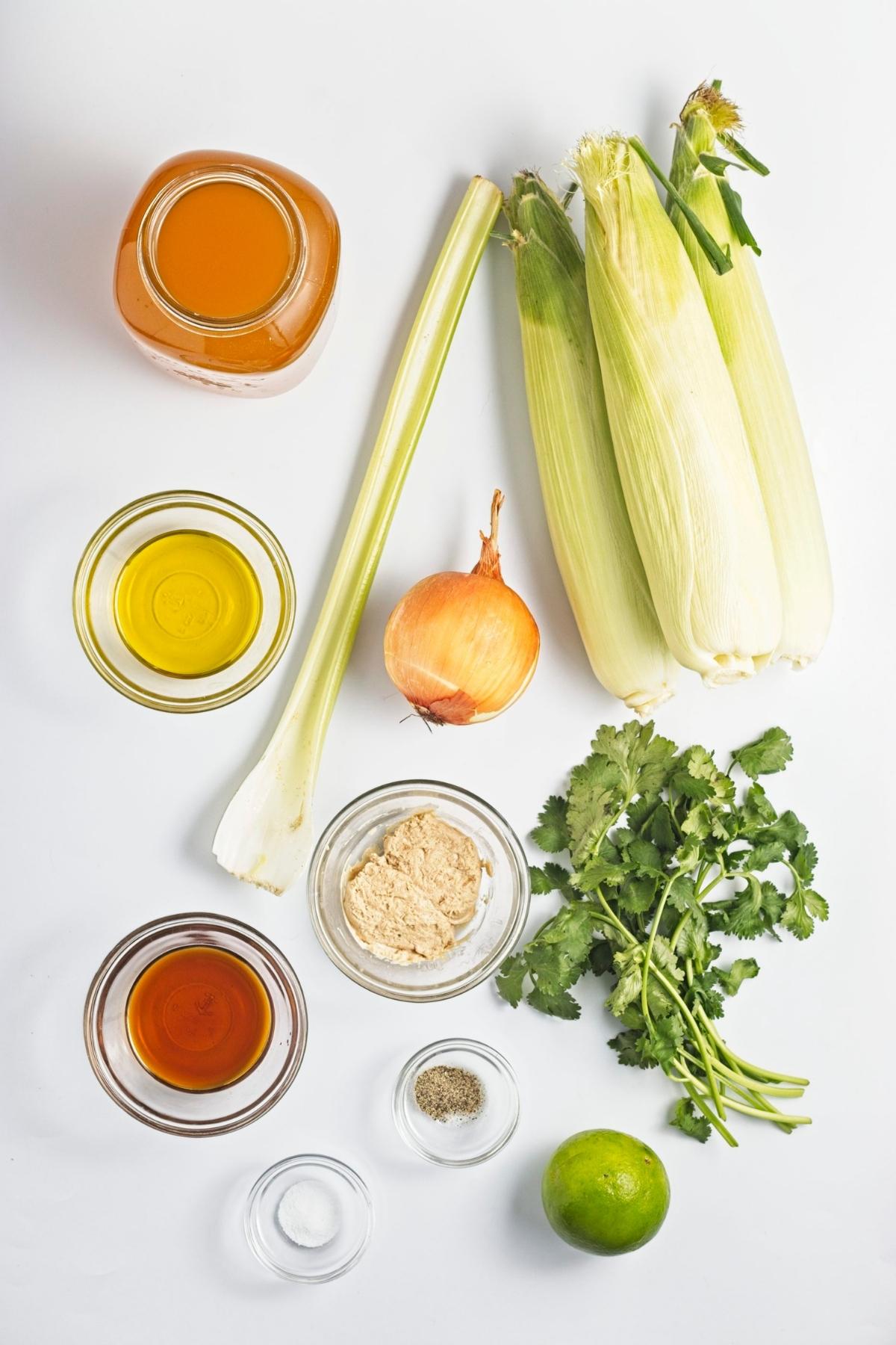 Ingredients arranged on a white surface including corn on the cob, onion, celery, miso paste, oil, broth, lime, and herbs.