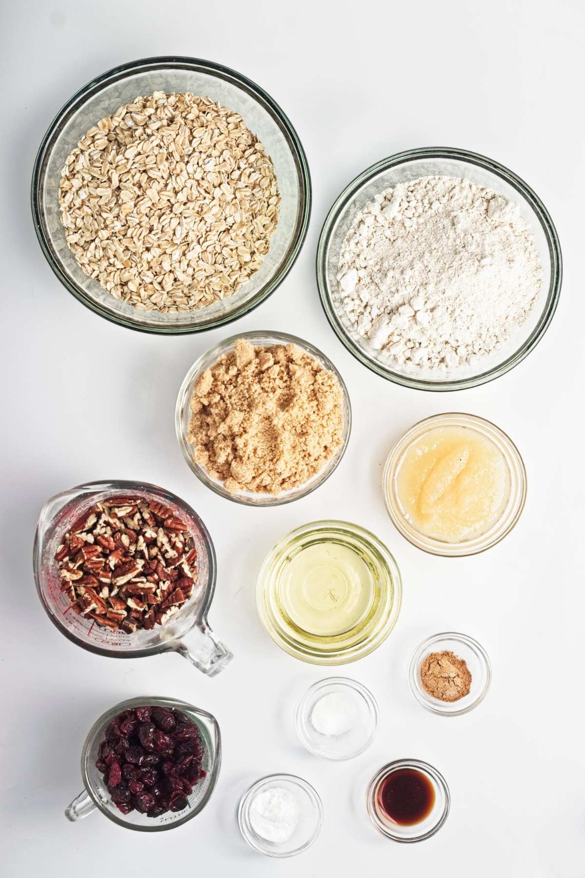 Measured bowls of oats, oat flour, brown sugar, oil, applesauce, pecans, cranberries, and spices on a white surface.