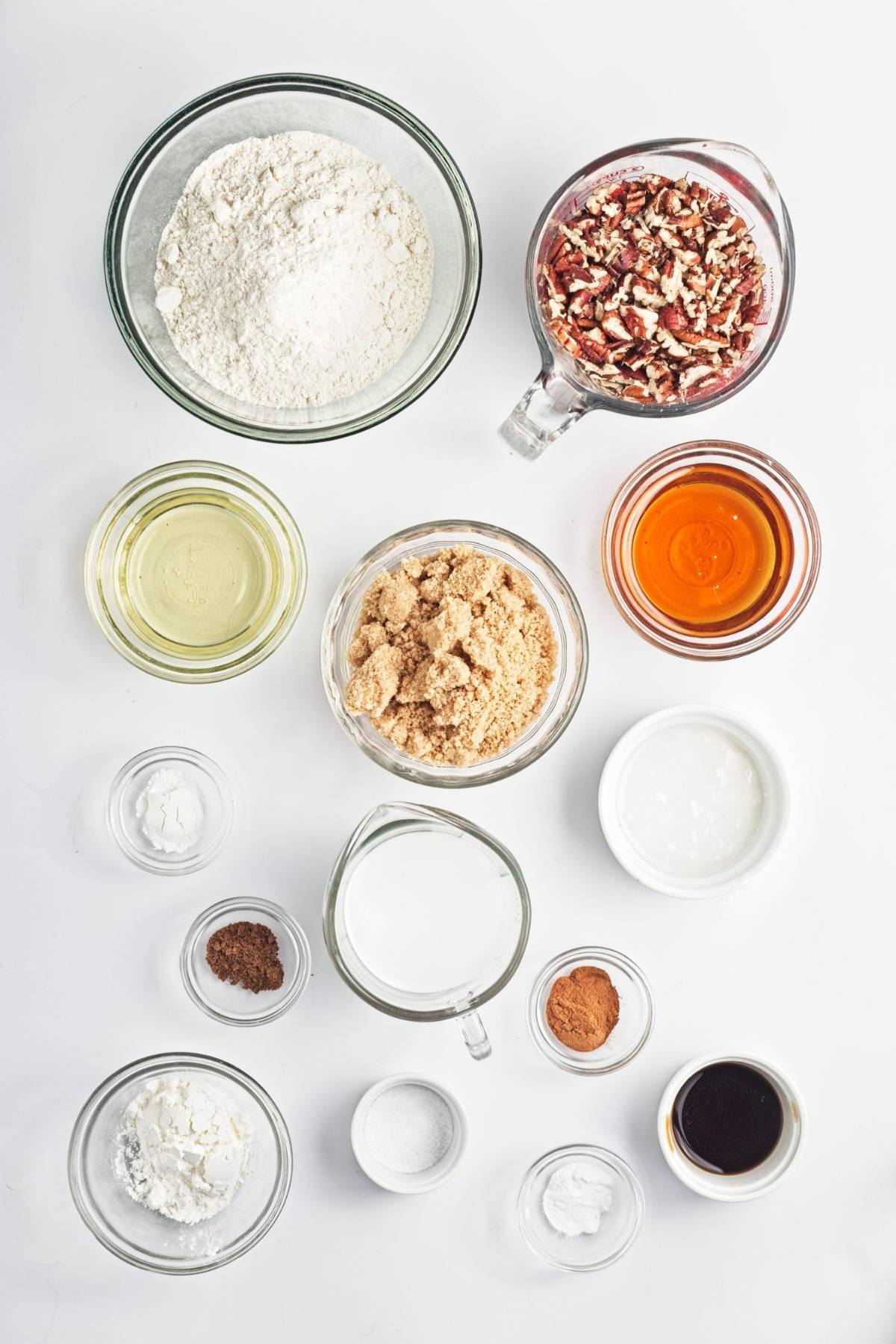 Flat lay of flour, chopped pecans, brown sugar, maple syrup, coconut milk, oil, vanilla, and spices in bowls.