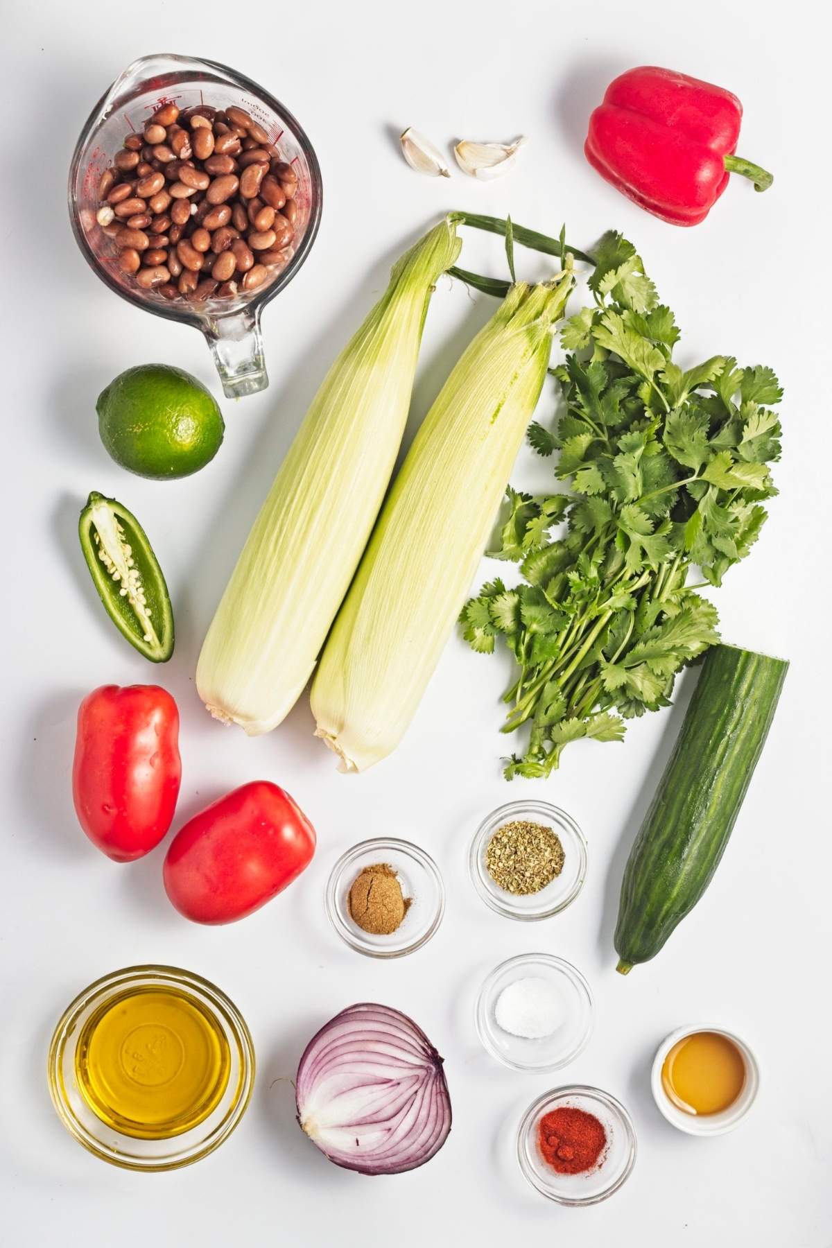 Fresh corn cobs, pinto beans, tomatoes, cucumber, red bell pepper, red onion, cilantro, lime, jalapeรฑo, garlic, olive oil, and spices arranged on a white surface.