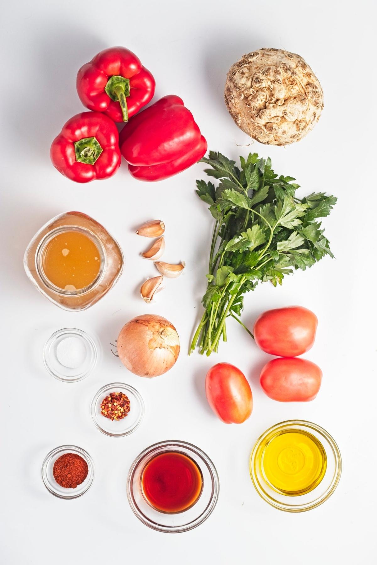 Flat lay of red bell peppers, tomatoes, onion, garlic, parsley, oil, vinegar, spices, and vegetable broth.