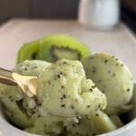 Kiwi sorbet scooped into a white bowl with visible kiwi seeds and sliced fresh kiwi behind.