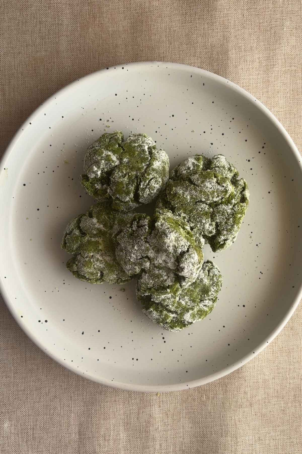 Top-down view of matcha crinkle cookies on a speckled plate, heavily coated in powdered sugar.