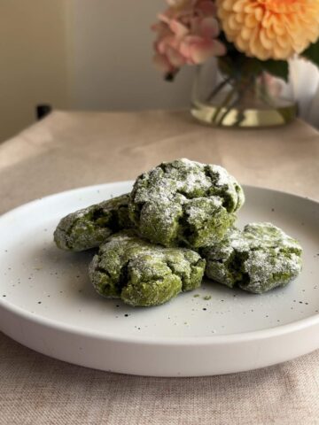 Stack of matcha crinkle cookies on a white plate, thick cracks and powdered sugar coating visible.