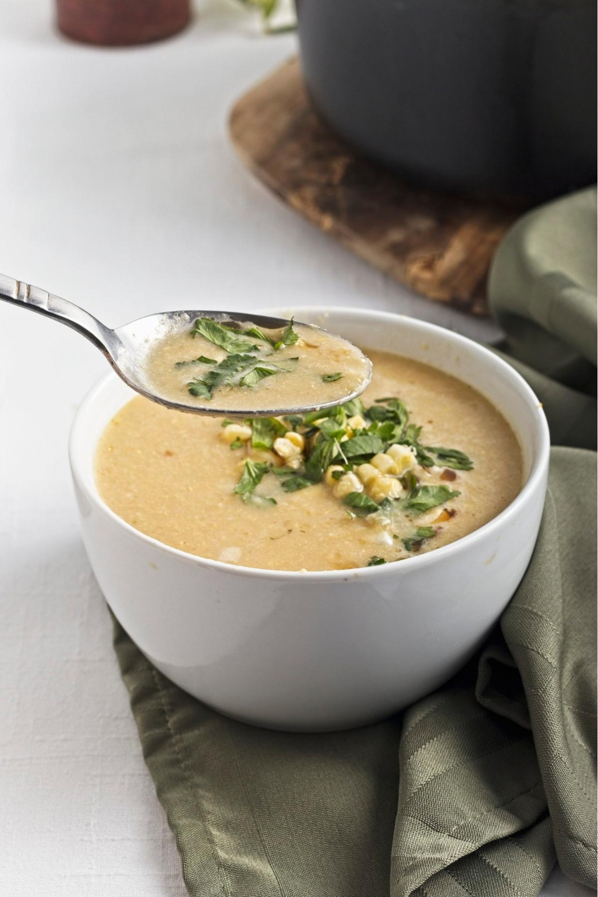Spoon lifting creamy corn soup from a white bowl.