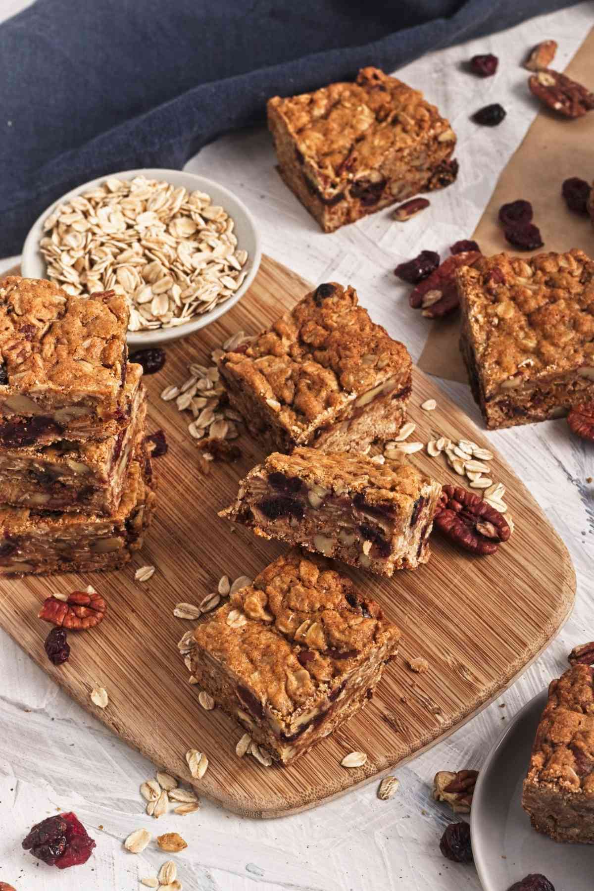 Oatmeal cookie bars arranged on a wooden board with pecans, dried cranberries, and rolled oats.