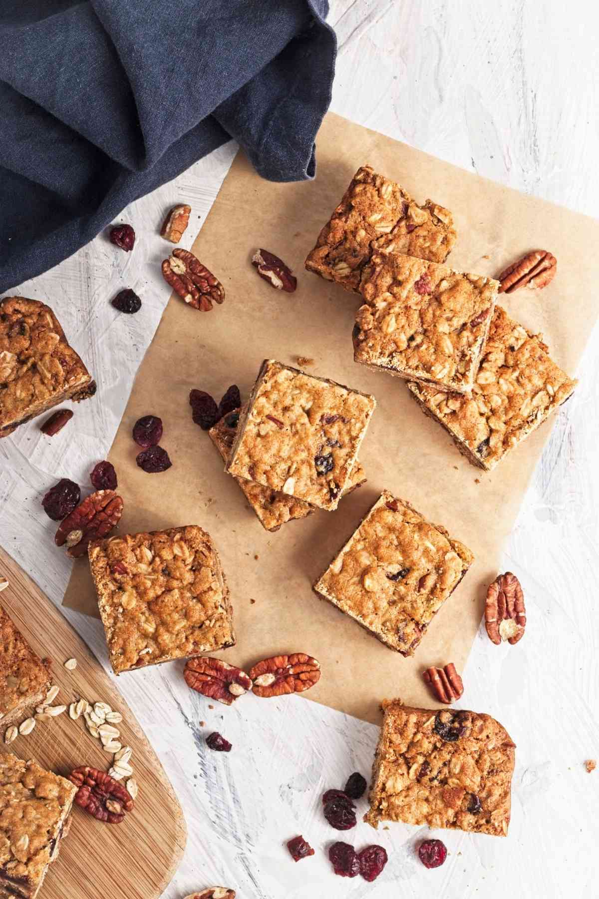 Stacked oatmeal cookie bars with cranberries and pecans on parchment, scattered oats and nuts around.