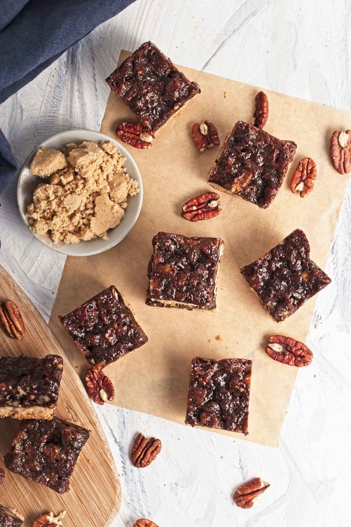 Cut pecan bars arranged on parchment with scattered pecans and a bowl of brown sugar.
