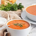 Two bowls of roasted red pepper soup served with bread and fresh herbs.