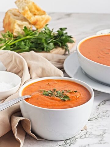 Two bowls of roasted red pepper soup served with bread and fresh herbs.